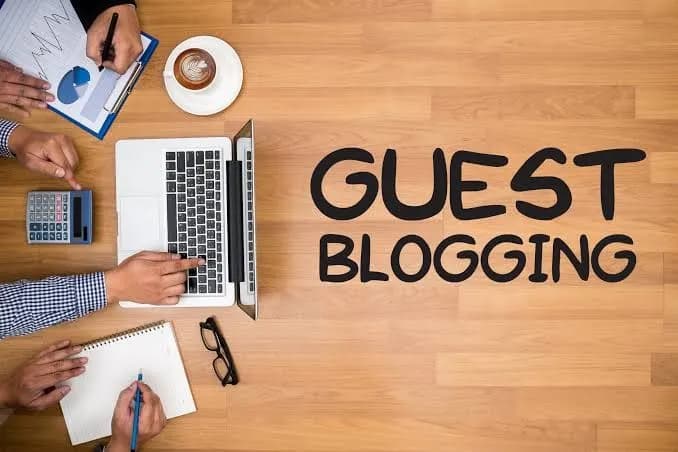 Overhead view of a laptop, notebook, calculator, and coffee cup beside the words "Guest Blogging" written on a wooden desk during a content planning session.